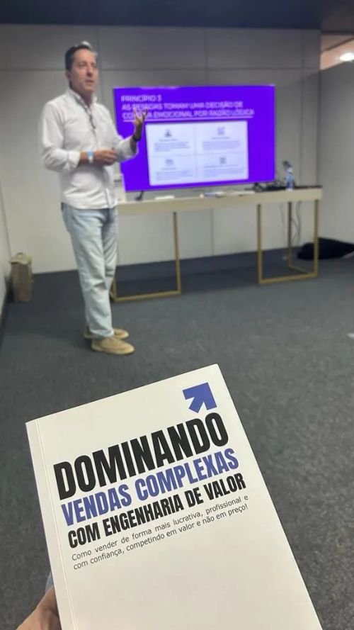 Em primeiro plano: Livro Dominando Vendas Complexas. Ao fundo: Marcos Mylius palestrando